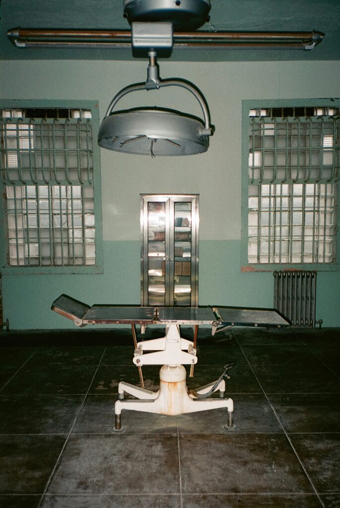 Abandoned operating room with a worn surgical table under a large overhead lamp, barred windows, and metal cabinets against pale green walls.